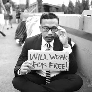 Un uomo seduto per terra con un cartello di cartone con scritto: I will work for free